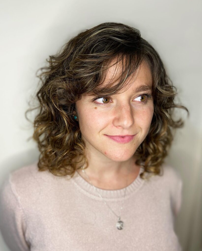 Shoulder length curly lob with soft layers and side swept bangs with subtle highlights.
