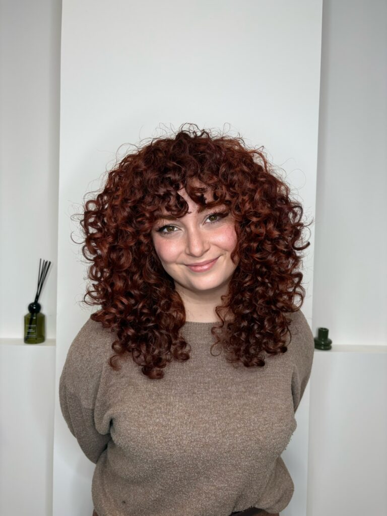 Rounded auburn curly hairstyle with full curly bangs and defined texture.