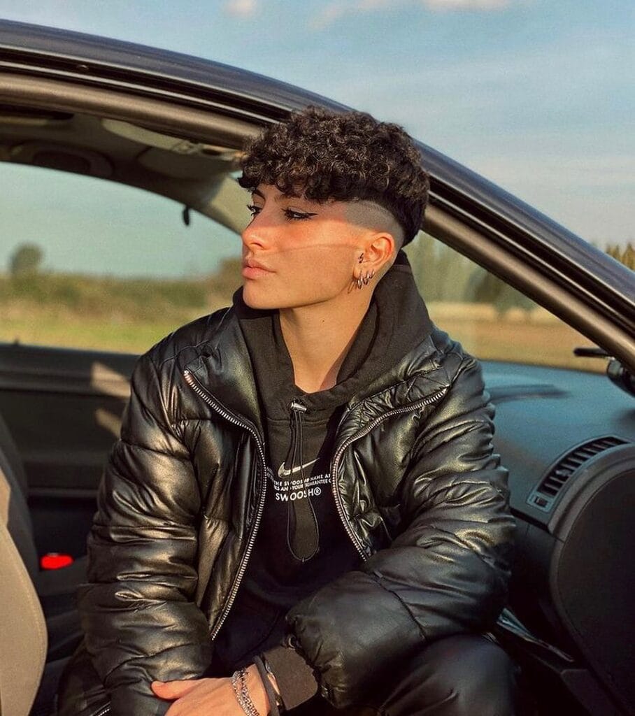 A short tomboy haircut with shaved sides and a curly textured top in natural dark hair.