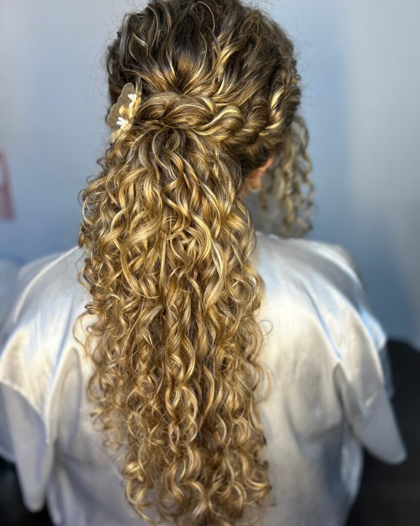 Long highlighted curly hair styled in a ponytail with braided crown details and defined curls.
