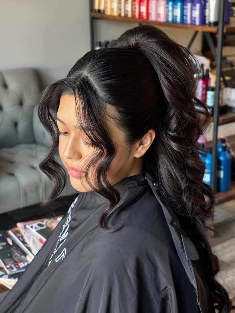 Long brown curly hair styled in a high voluminous ponytail with a braided crown and loose face framing curls.