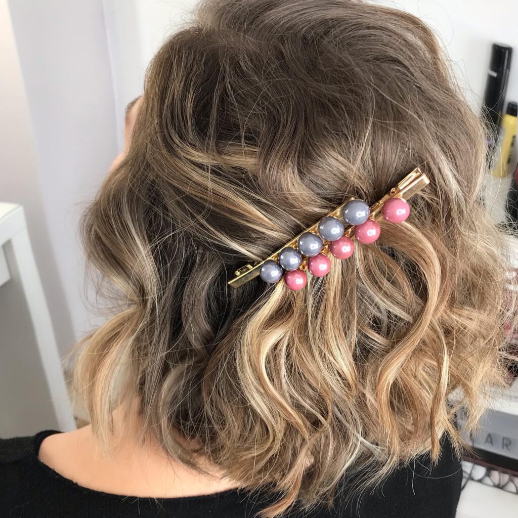 A woman with a curly balayage bob wears a diagonal pink and grey beaded clip pinning back the top section.