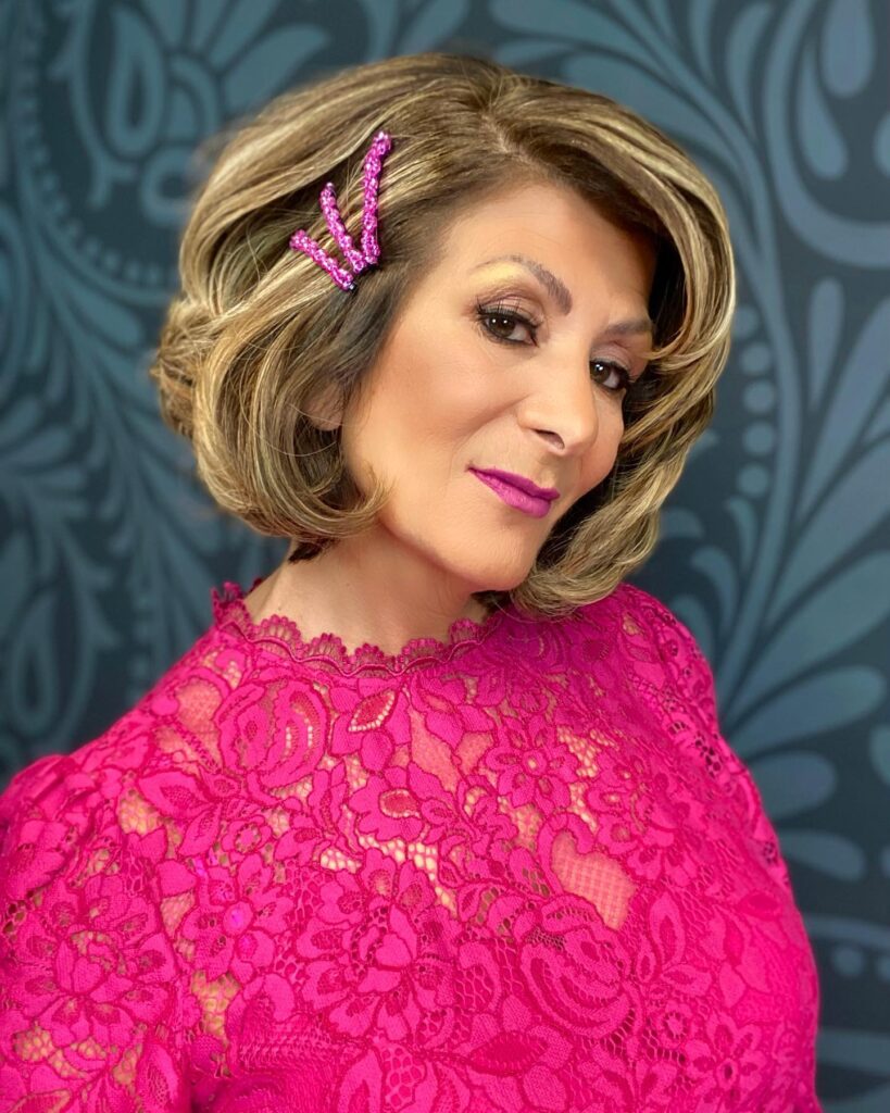 A woman with a voluminous curly blonde and brunette bob wears a sparkly pink crystal hair clip on one side.