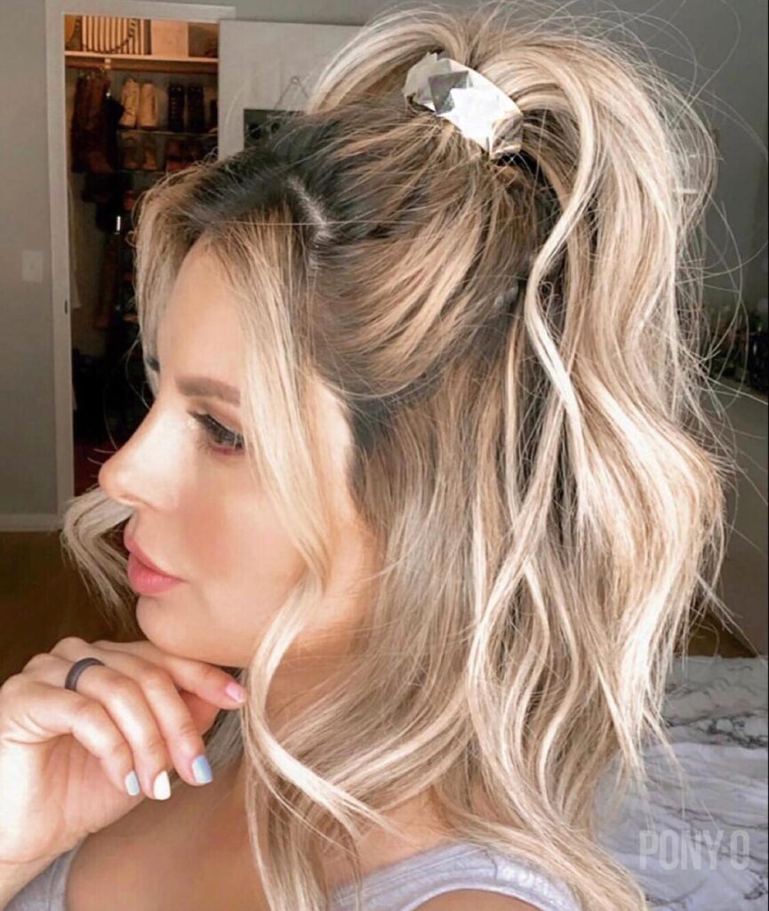 A woman with wavy blonde balayage lob length hair wears a small claw clip gathering the top section high on her crown.