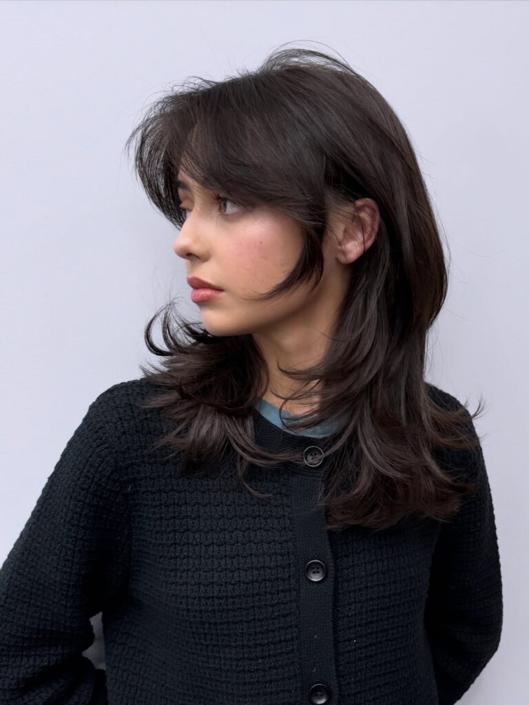 A woman with a shoulder-length brunette shag haircut featuring feathery curtain bangs and choppy face-framing layers.
