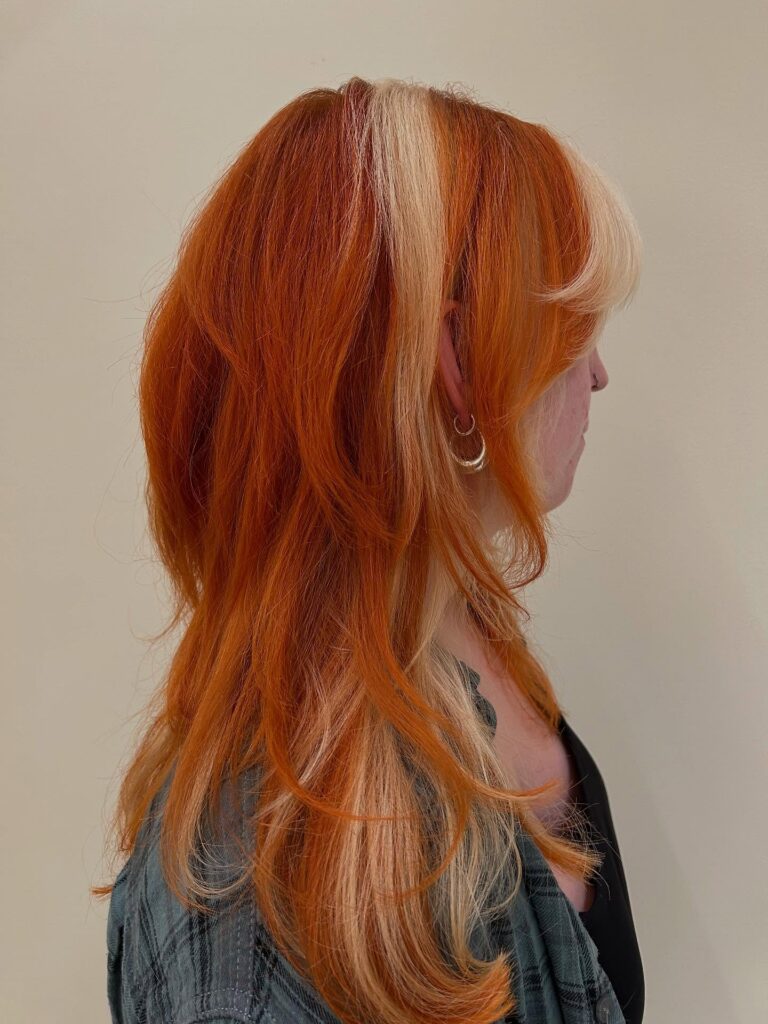 A woman with long layered copper orange hair featuring a chunky blonde money piece.