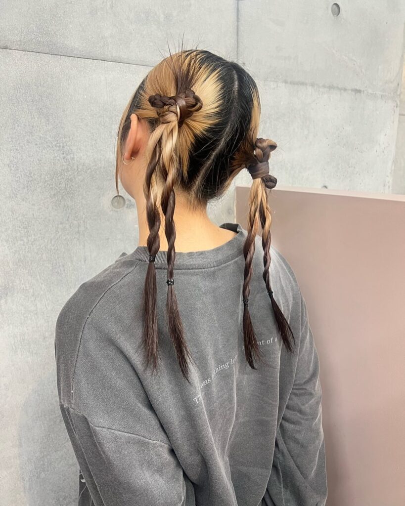 A woman with dark brown hair featuring blonde money pieces styled into four small knotted braids at the back.