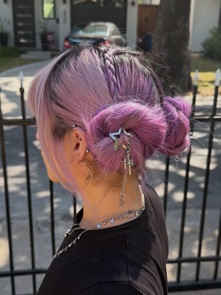 A woman with pastel lavender and pink hair styled into two low buns with a center braid and a star charm clip.