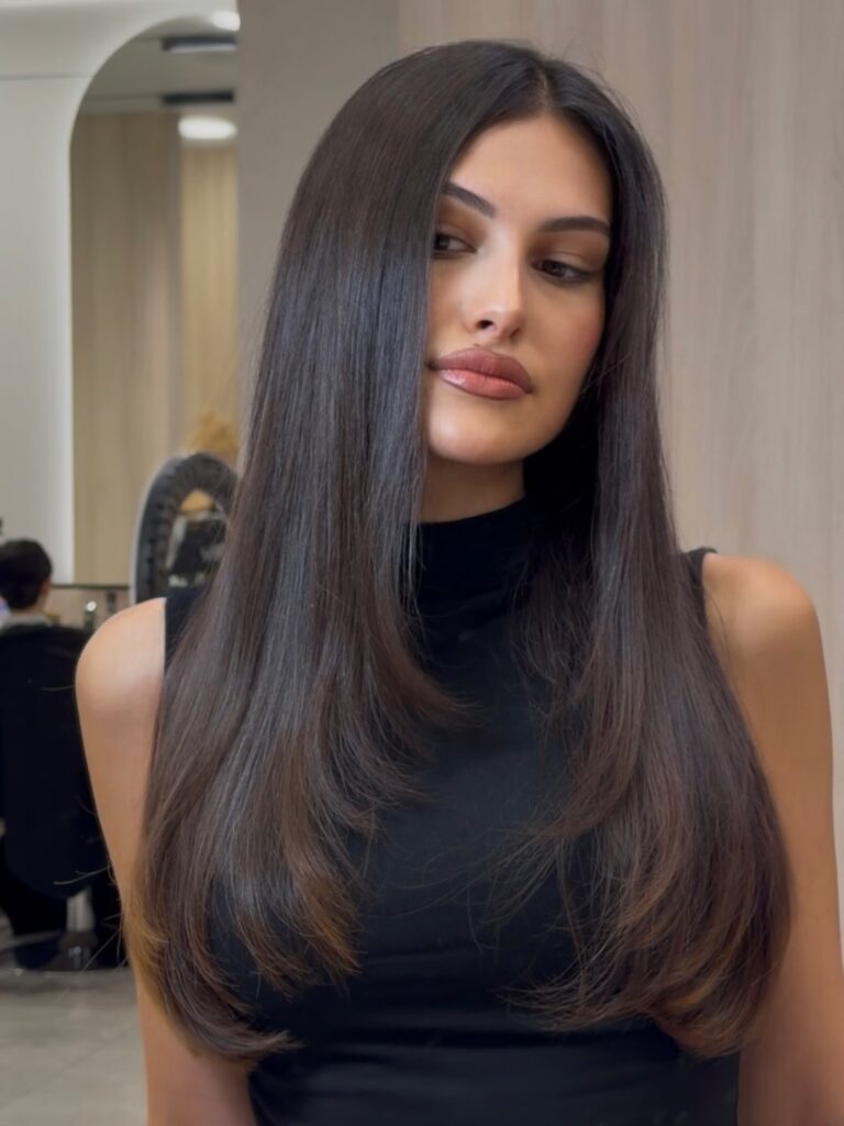 A woman with long, sleek, straight dark brown hair and subtle caramel highlights wearing a black top indoors.