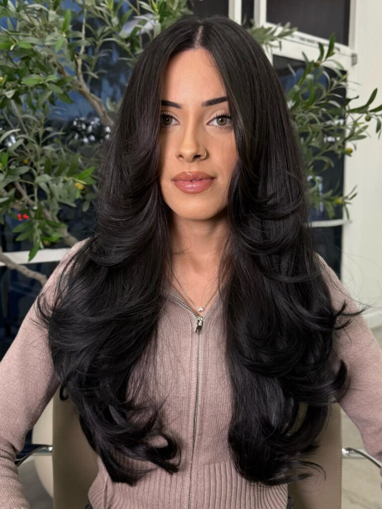 A woman with long, voluminous black hair styled in bouncy curtain layers standing in front of greenery.