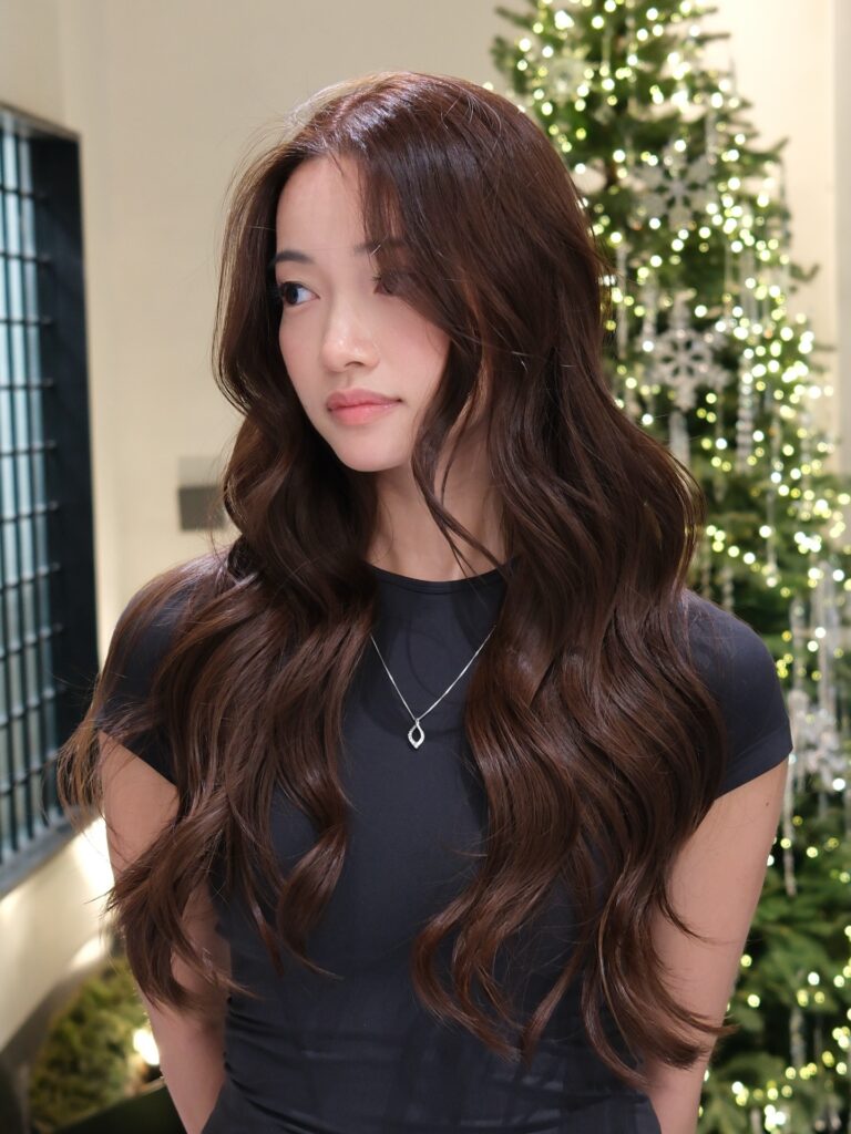 A woman with long, wavy chocolate brown hair and wispy curtain bangs standing in front of a lit Christmas tree.