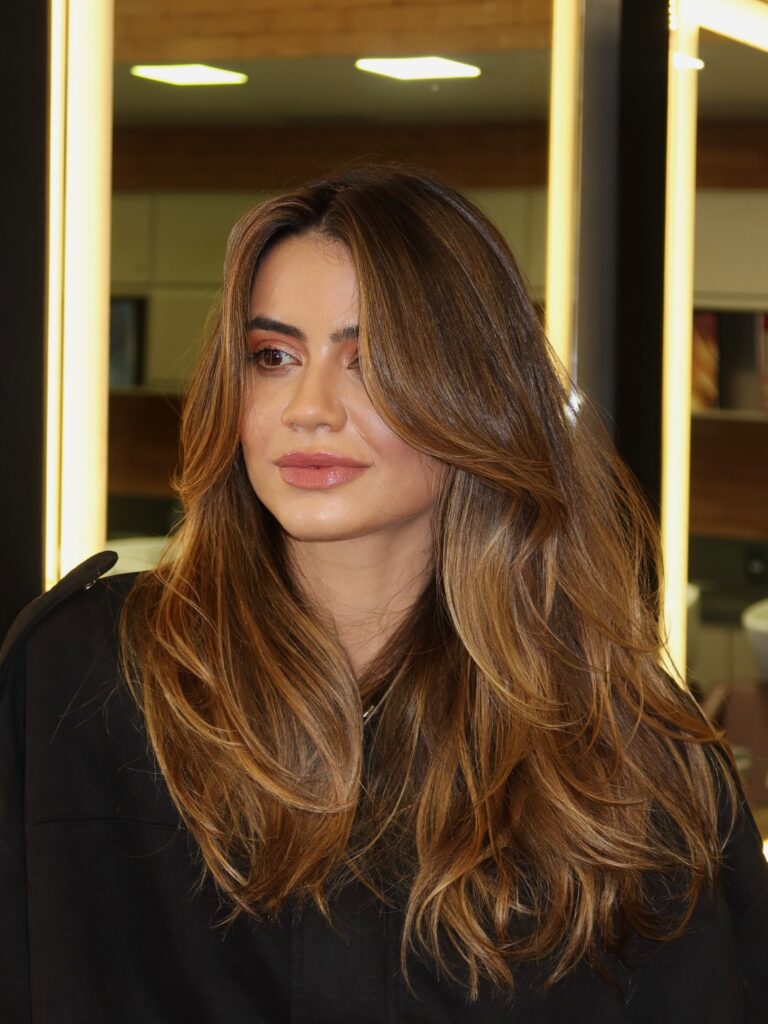 Woman with long warm chestnut hair featuring caramel balayage highlights and voluminous center parted curtain bangs.