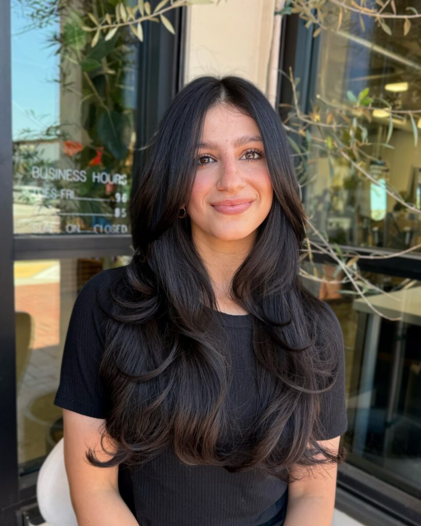 A woman with long, wavy, layered black hair and center-parted curtain bangs smiling outside a storefront.