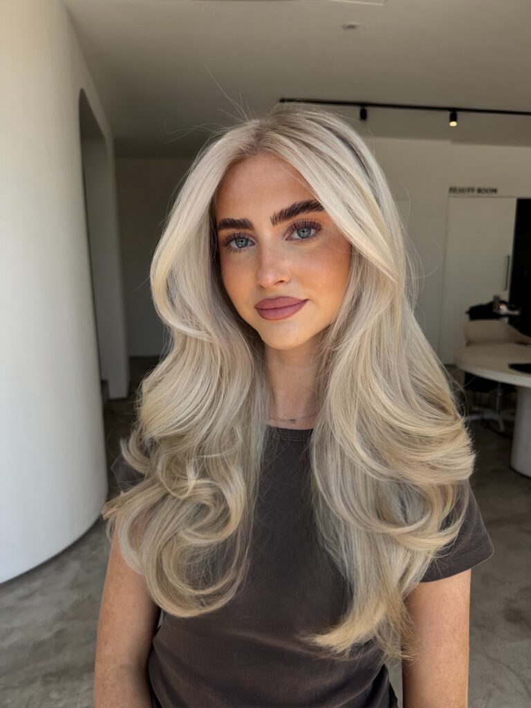Woman with long bright platinum blonde layered hair styled in bouncy curls with center parted curtain bangs.