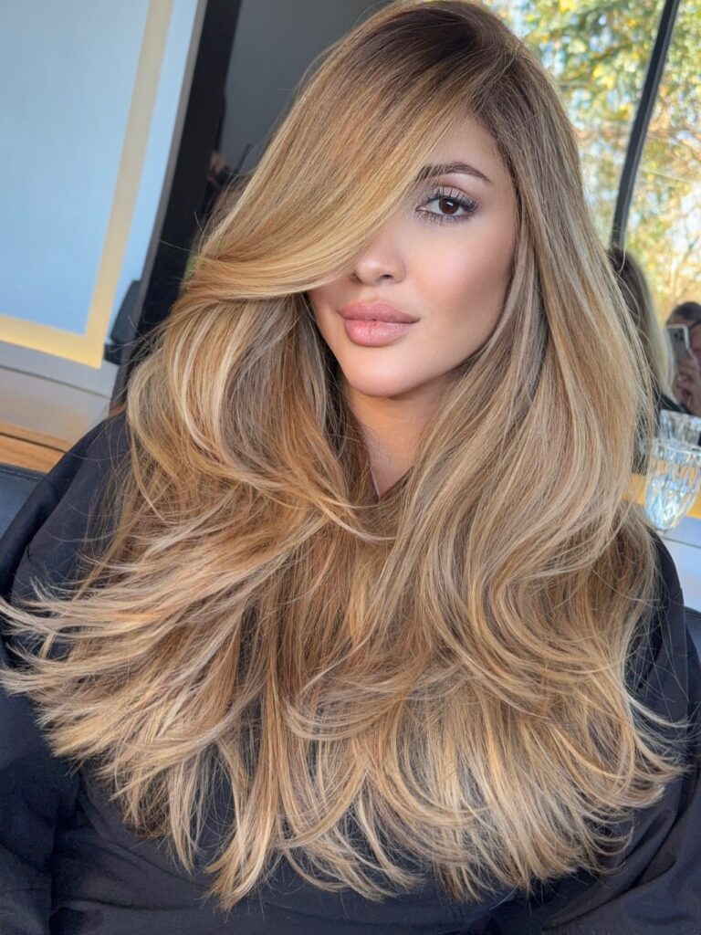 A woman with long, golden balayage waves and a deep side-swept fringe posing outdoors.