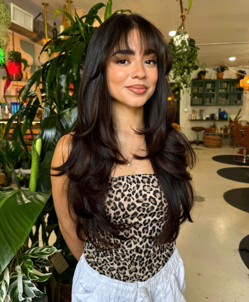 Woman with long glossy dark chocolate brunette hair styled with layered ends and wispy curtain bangs.