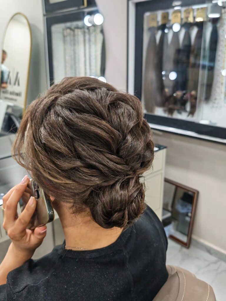 A woman with brunette hair wears an intricate low braided updo with twisted sections gathered into a bun at the nape of her neck.