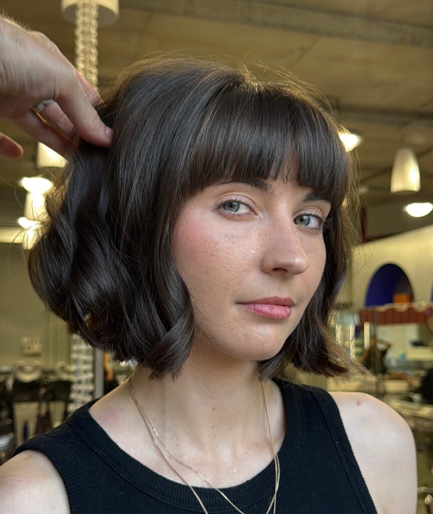 A woman with a dark brunette wavy bob and full blunt bangs poses for a close up portrait.