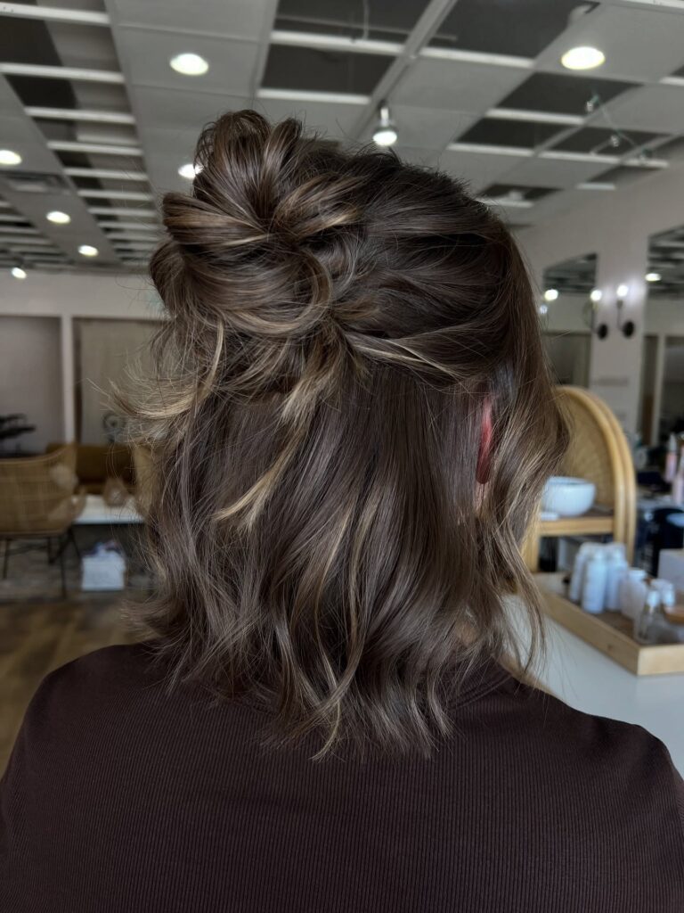 A woman with brunette hair featuring caramel highlights wears a messy half-up twisted bun with loose wavy layers falling below her shoulders.