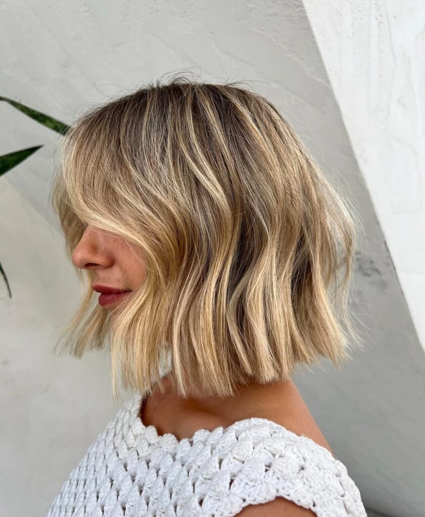 A woman with a textured golden blonde wavy bob and curtain bangs is shown in profile.