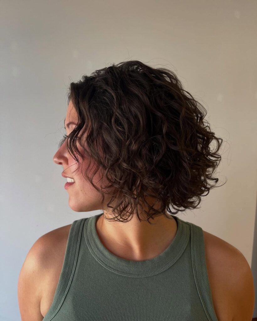 A woman with a voluminous chin-length curly brunette bob showing bouncy spiral texture from the side.