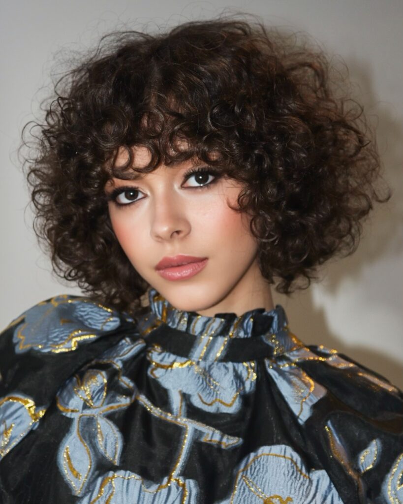 A woman with a chin-length dark brunette curly bob styled with a short piecey fringe and full round shape.