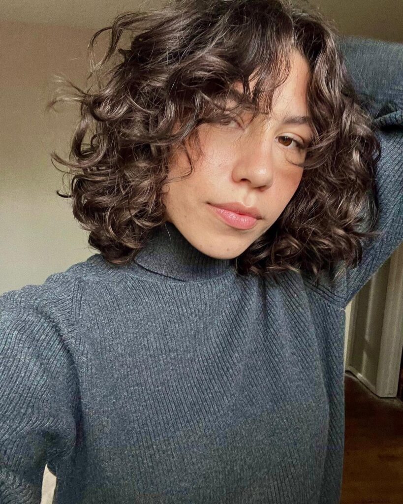 A woman with a shoulder-length curly brunette lob featuring caramel highlights and face-framing ringlets.