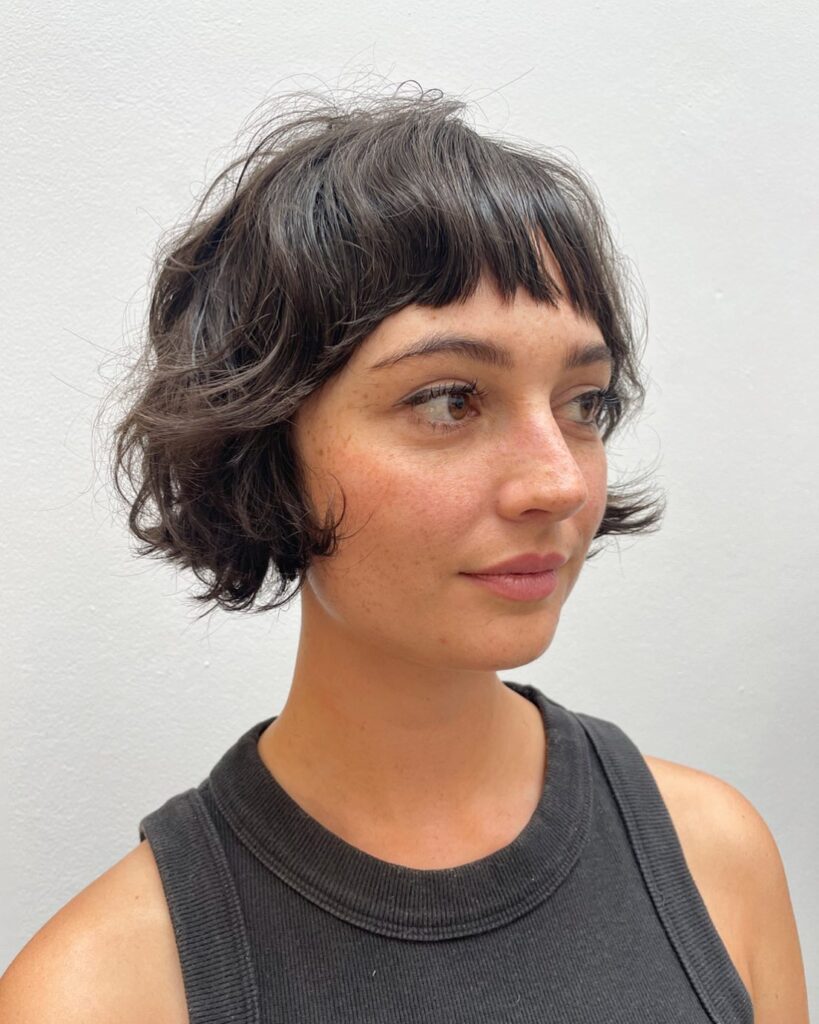 A woman with a chin-length curly brunette bob and wispy fringe styled with tousled, face-framing waves.