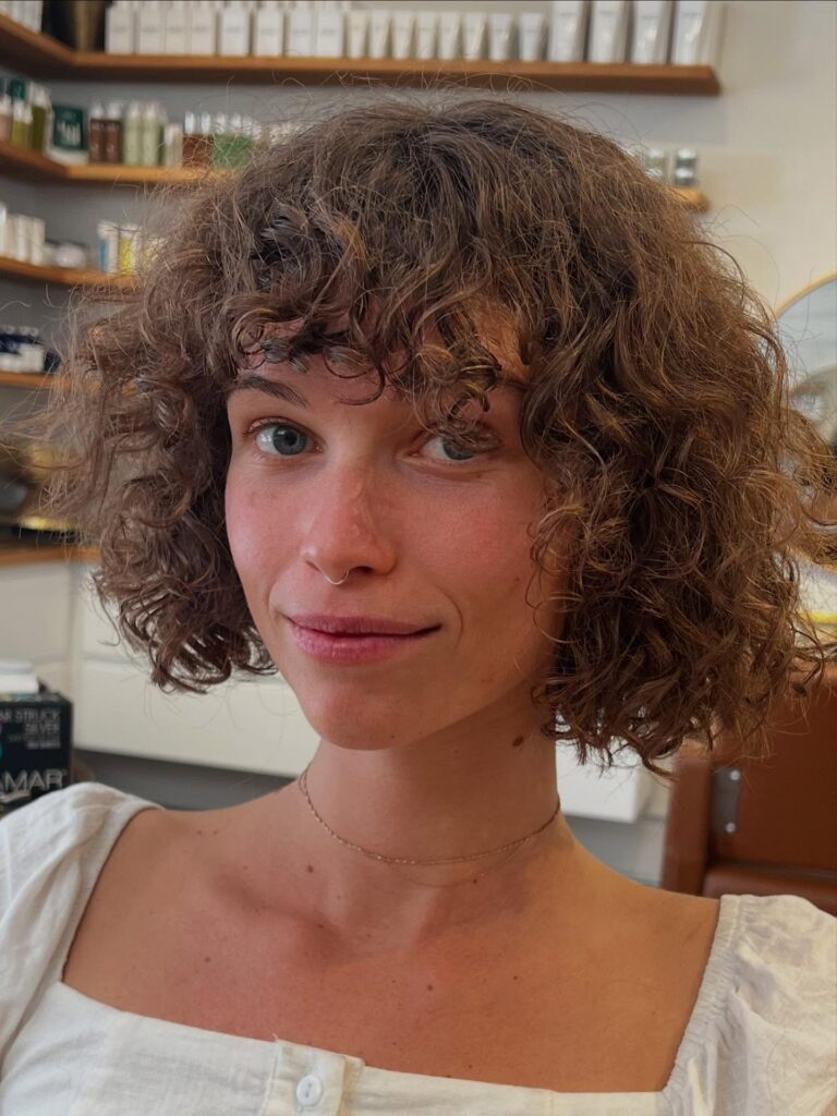 A woman with a chin-length curly brown bob styled with a full curly fringe that blends into the layers.