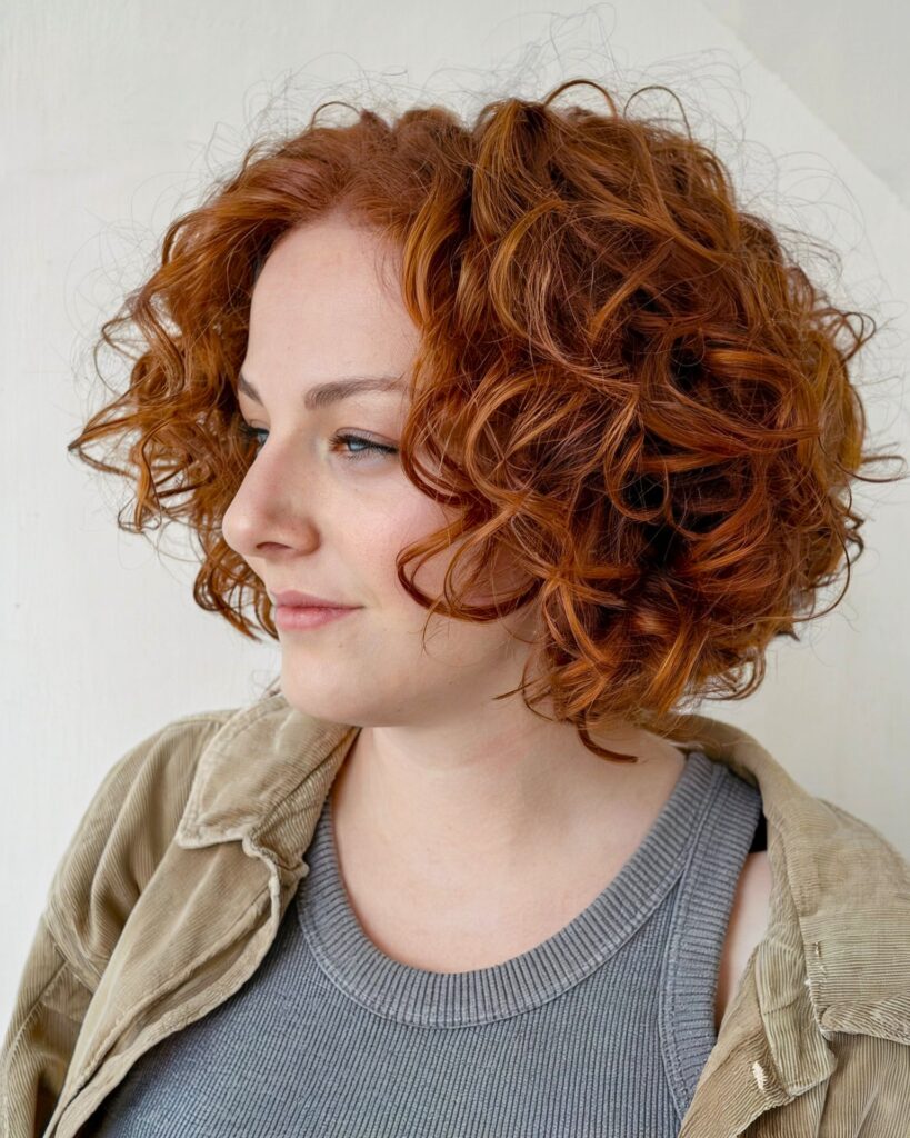 A woman with a chin-length curly ginger red bob styled with a deep side part and full round volume.