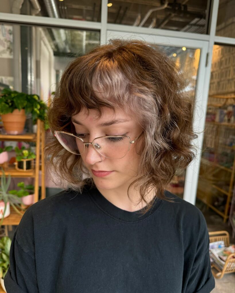 A woman with a shoulder-length curly brown wolf cut featuring money piece highlights and wispy curtain bangs.