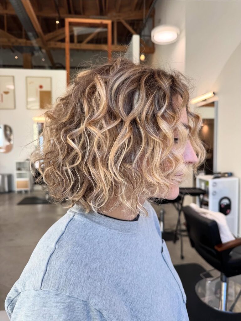 A woman with a chin-length curly balayage bob styled with tousled beachy texture and blonde highlights.