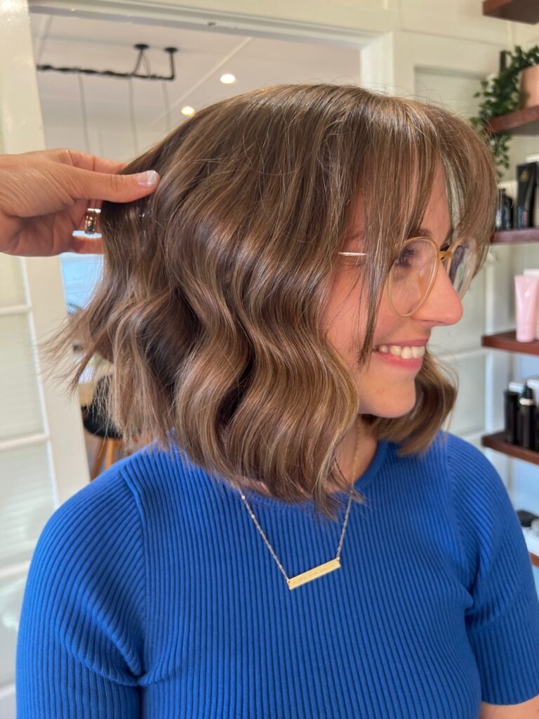 A woman with a wavy balayage lob and curtain bangs smiles while someone lifts a section of her hair.