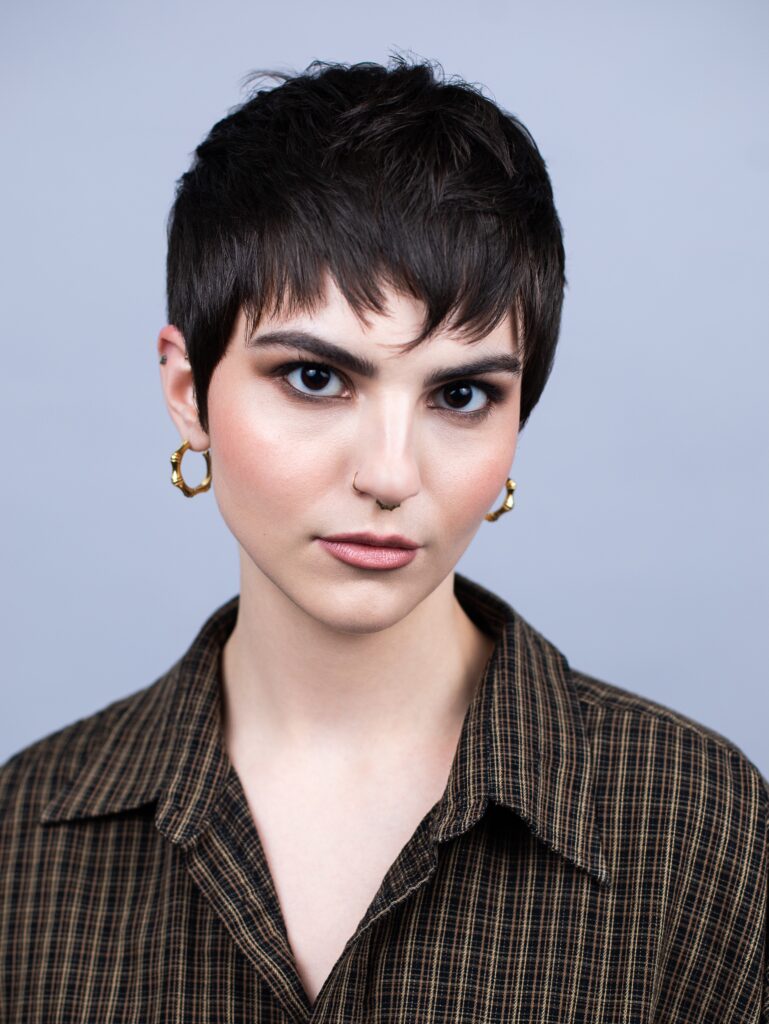 A woman with a black cropped pixie cut and choppy fringe poses in a plaid shirt.
