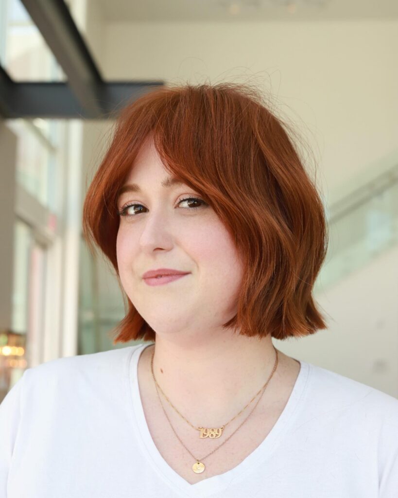 A woman with a wavy copper red bob and curtain bangs smiles gently at the camera.