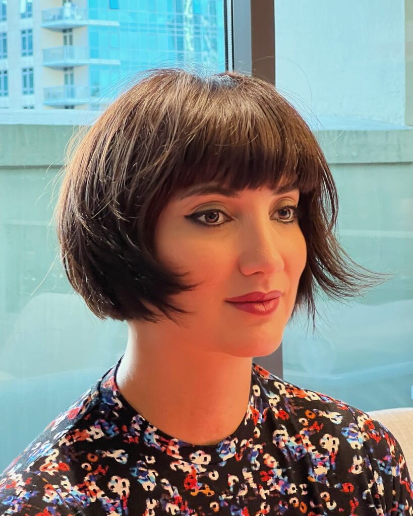 A woman with a chocolate brunette chin-length bob and blunt micro bangs stands in front of a window.