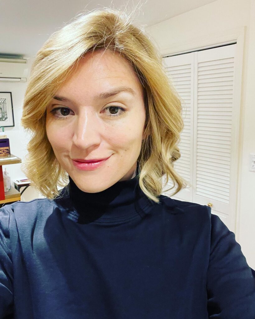 A woman with a golden blonde bob and bouncy curled ends takes a selfie smiling.