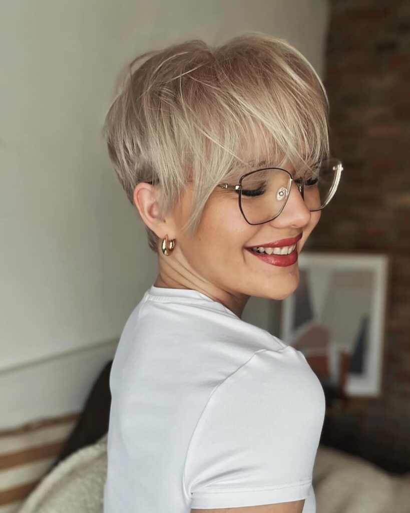 A woman with a platinum blonde pixie cut and blunt fringe smiles while wearing glasses.