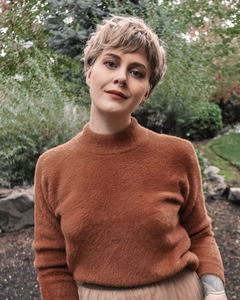 A woman with a honey blonde pixie cut and wispy fringe smiles softly outdoors.