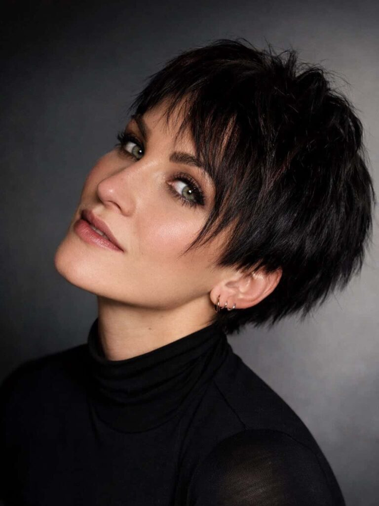 A woman with a black textured shaggy pixie cut and face framing fringe tilts her head to the side.