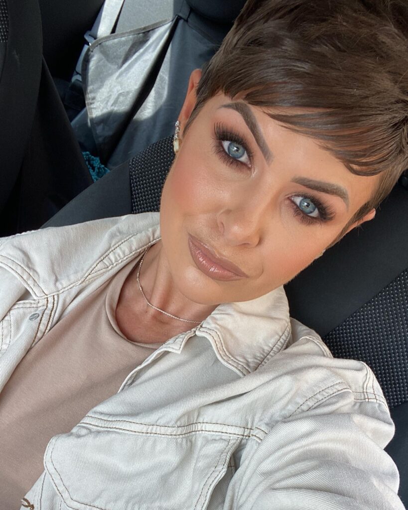 A woman with an espresso brown pixie cut and long side swept bangs takes a selfie in a car.
