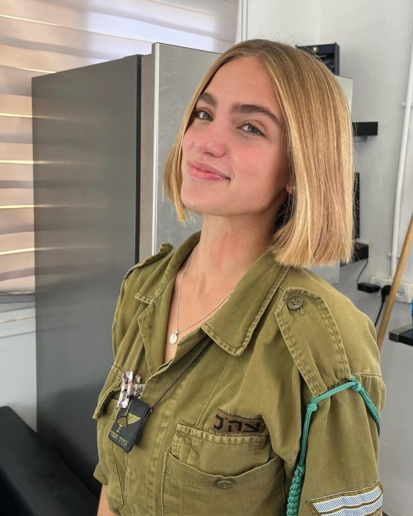 A woman with a straight golden blonde lob and side part wears a green utility jacket with a necklace.