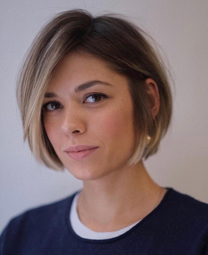 A woman with a jaw-length balayage bob and deep side part wears a navy and white shirt.