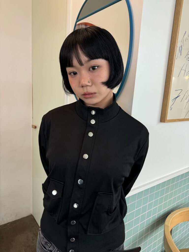 A woman with a sleek black chin-length bob and micro bangs wears a black snap-button jacket in front of a round mirror.