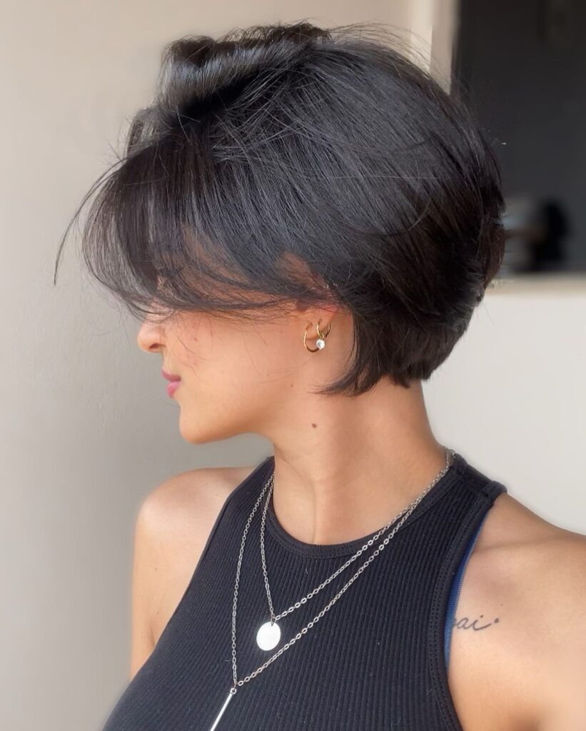 A woman with a voluminous dark pixie haircut and long side fringe wears layered silver necklaces and hoop earrings.