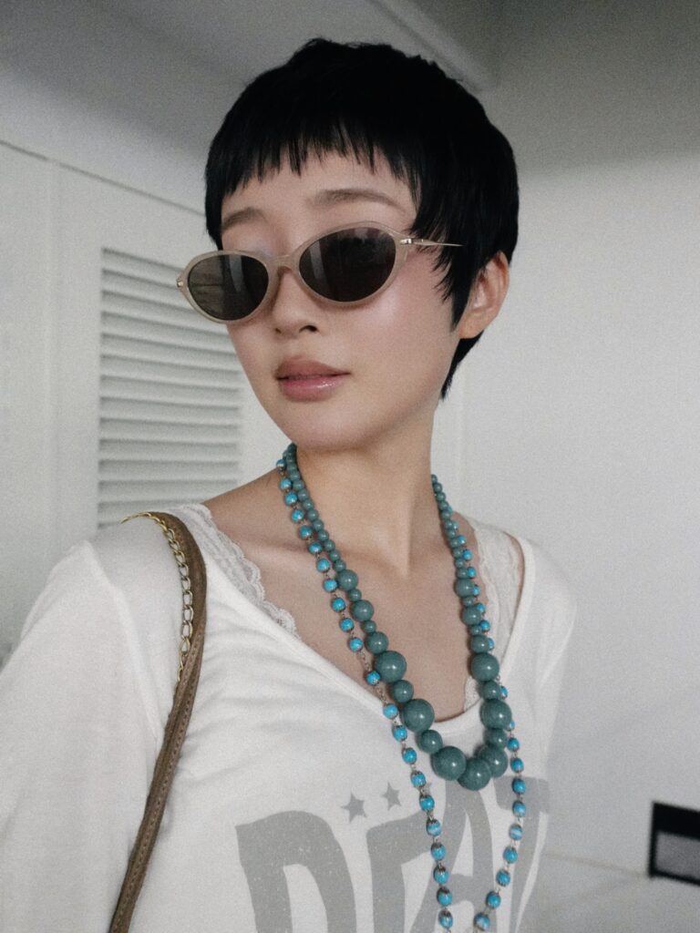 A woman with a short black textured pixie haircut and blunt bangs wears round sunglasses and layered turquoise beaded necklaces.