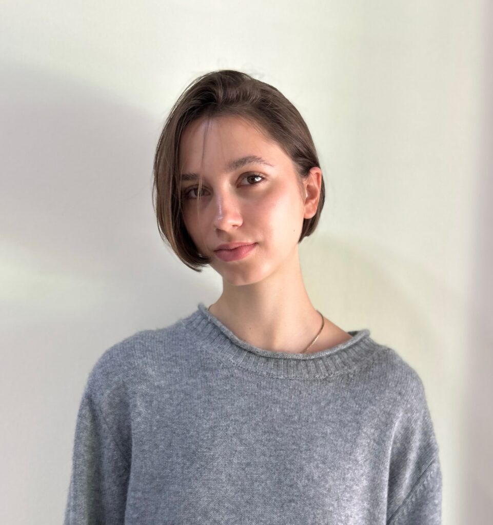 A woman with a center-parted brunette chin-length bob wears a grey knit sweater and a delicate necklace.