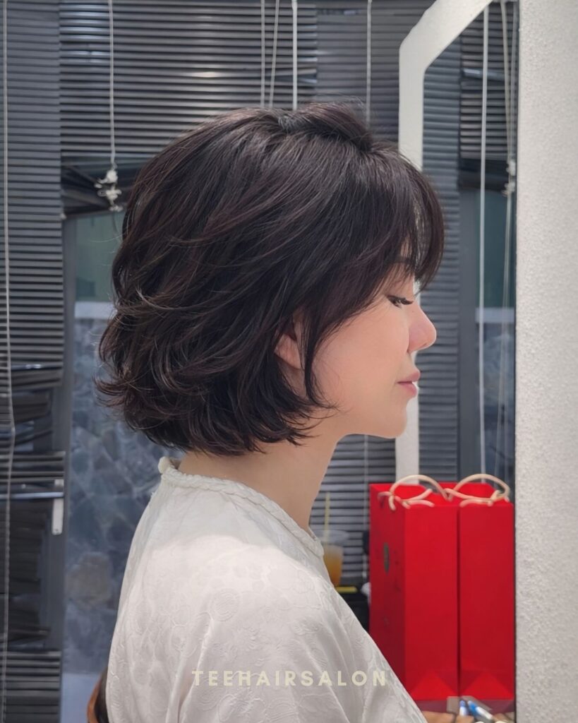 A woman with a flipped-out layered brunette bob and wispy bangs is photographed in profile against striped blinds.