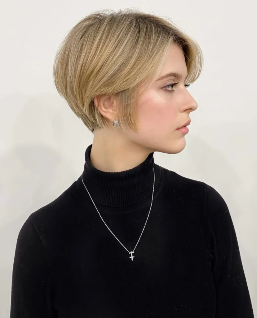 A woman with a sleek honey blonde pixie and long sweeping bangs wears a black turtleneck and a cross necklace.