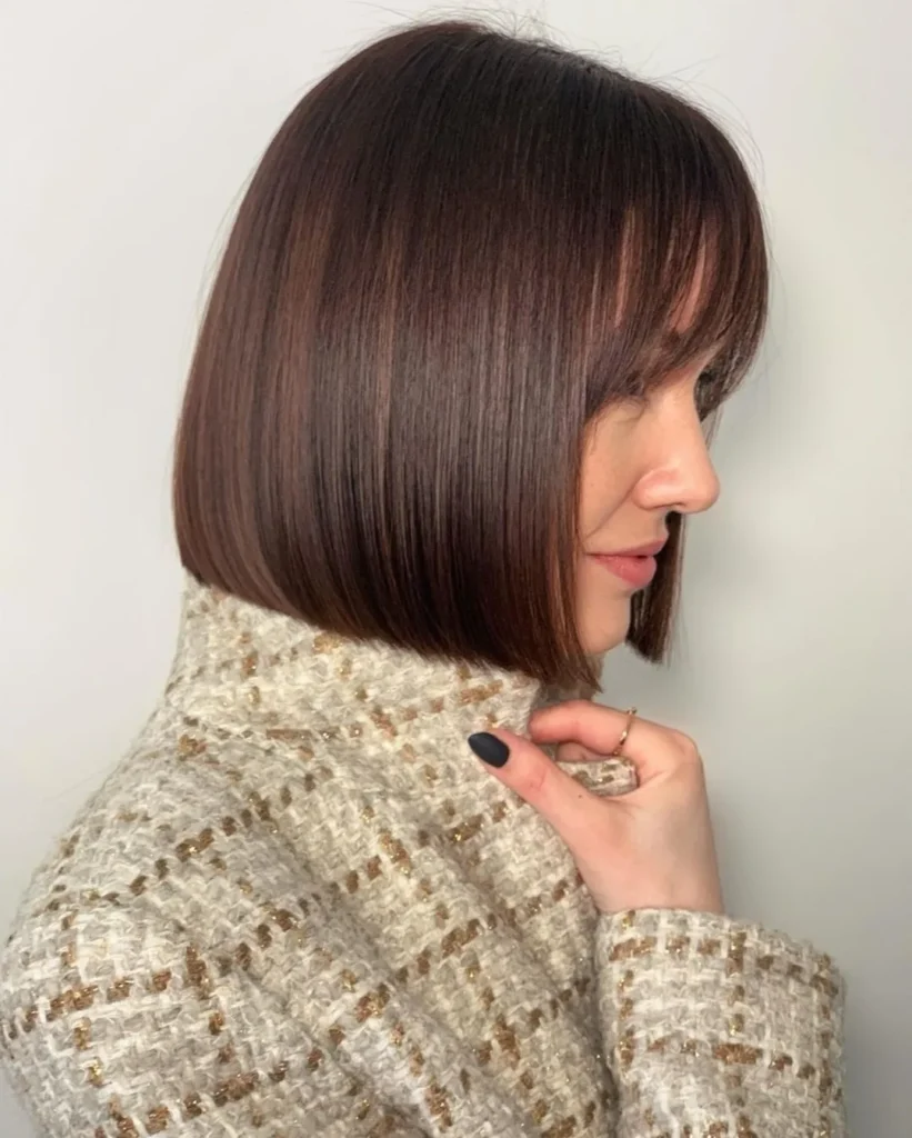 A woman with a glossy auburn blunt bob and wispy bangs wears a tweed jacket with gold rings.